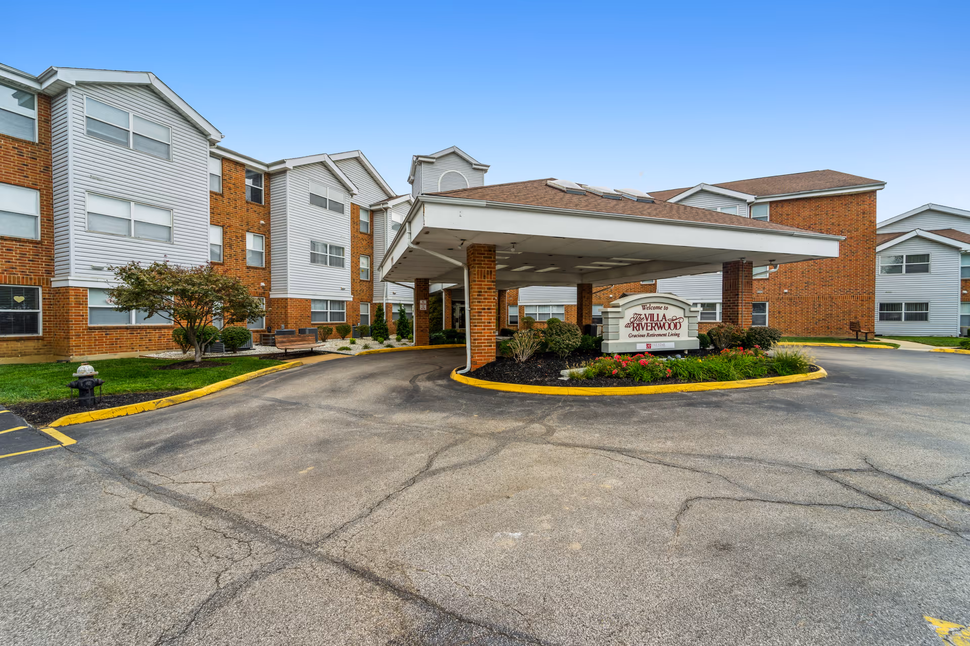 Front entrance canopy and driveway of The Villa at Riverwood senior living facility with brick-and-siding buildings and landscaping.