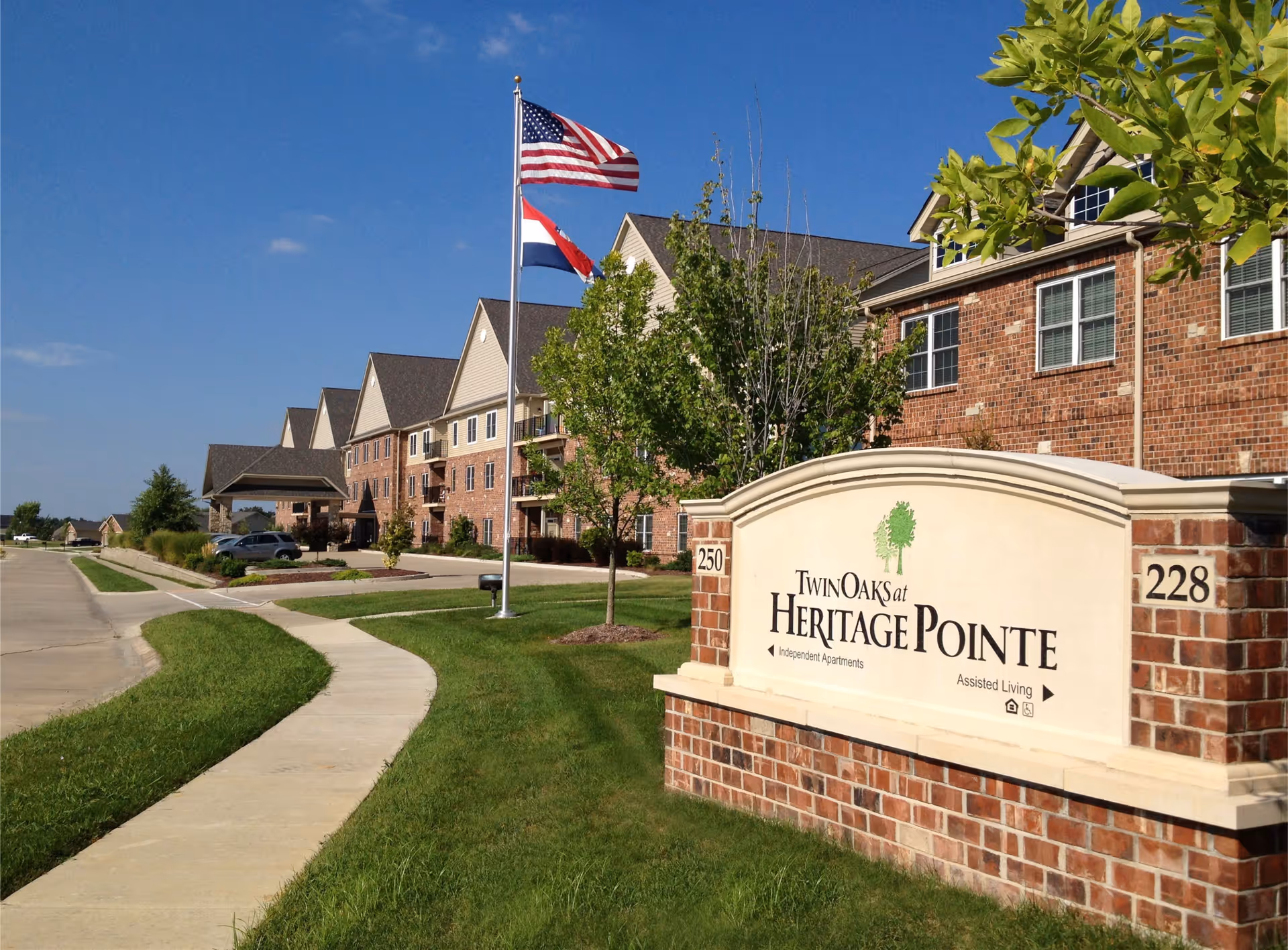 Exterior view of Twin Oaks at Heritage Pointe senior living facility with a brick building, a sidewalk, green grass, trees, and two flagpoles flying the American and state flags under a clear blue sky.