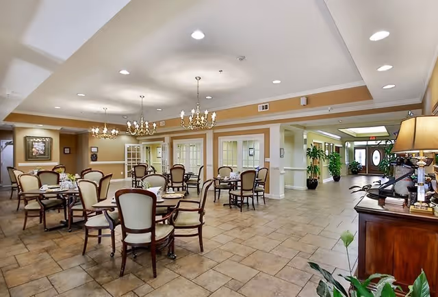 A spacious dining room in a senior living facility with multiple round tables and cushioned chairs arranged neatly. The room features beige walls with white trim, tiled floors, elegant chandeliers hanging from the ceiling, and large windows and glass doors in the background. There is a wooden sideboard with a lamp and some decorative items on the right side, and plants near the entrance area in the distance.