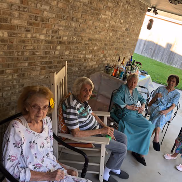 Four elderly women sitting on chairs on a covered patio with a brick wall behind them. They appear to be socializing and relaxing, with a table in the background holding various bottles and items. The patio opens to a grassy yard enclosed by a wooden fence.