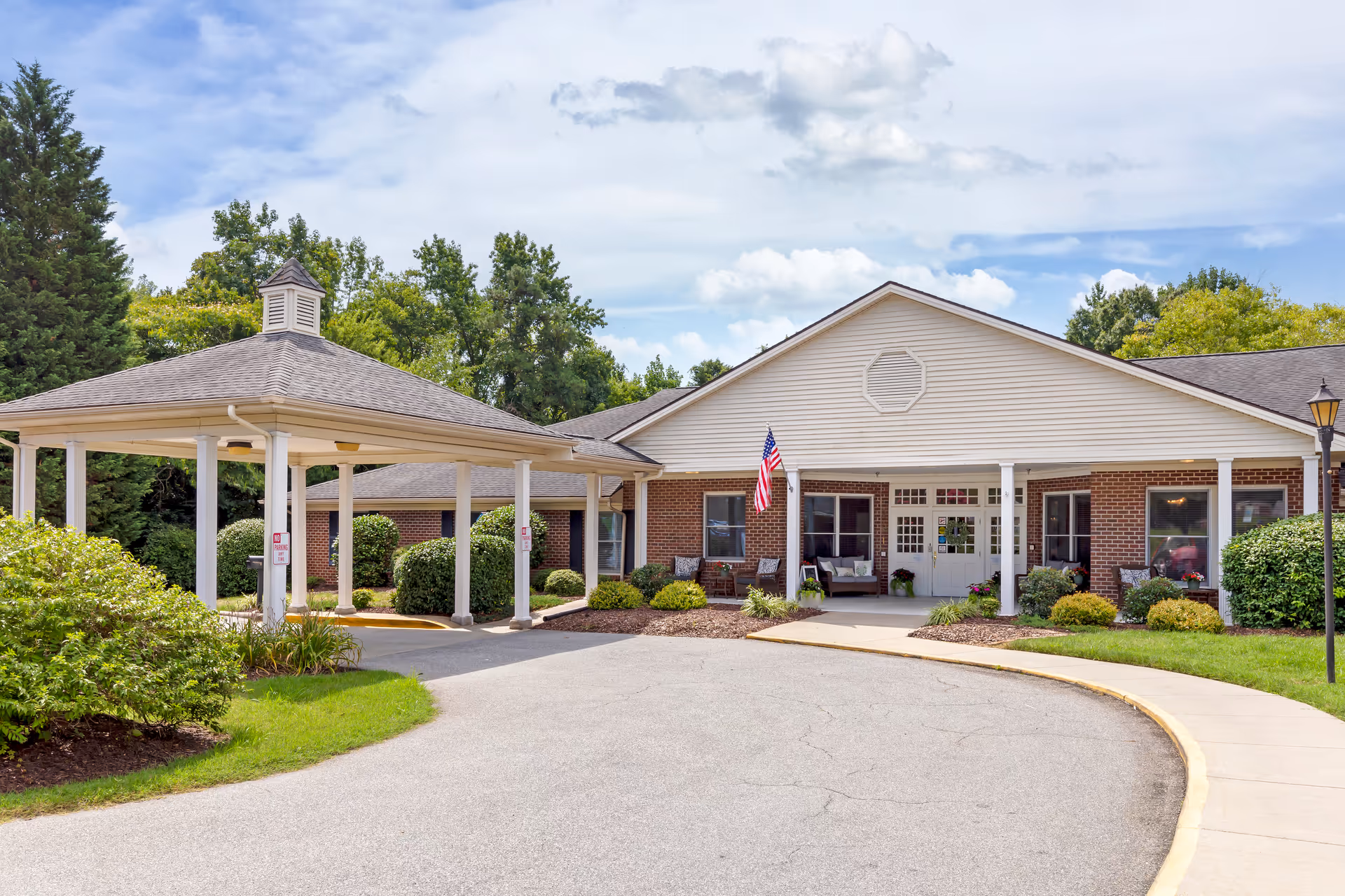 Front exterior view of a single-story brick building with a covered entrance driveway, white columns, and an American flag hanging near the entrance. The building is surrounded by green bushes, trees, and a well-maintained lawn under a partly cloudy sky.