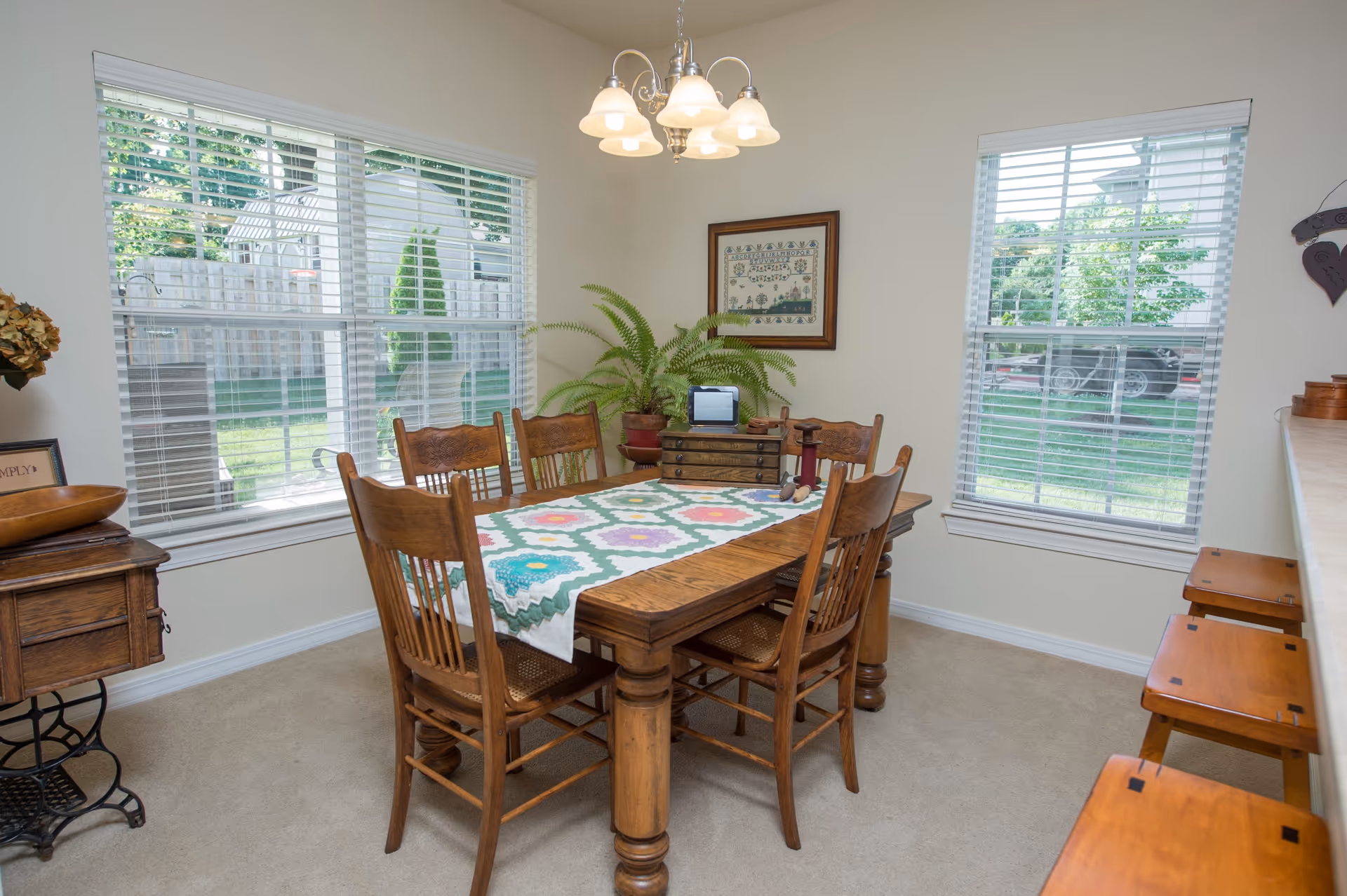A cozy dining room with a wooden table and six matching chairs. The table is decorated with a colorful quilted table runner and various small decorative items. Two large windows with white blinds let in natural light and show a green outdoor area. A framed picture hangs on the wall, and a hanging light fixture with five lamps illuminates the room. There are also three wooden stools lined up along a counter on the right side.