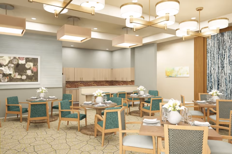 A well-lit dining room with multiple round and square wooden tables set with white plates, napkins, glassware, and white floral centerpieces. The chairs have wooden frames with teal and patterned upholstery. The room features modern ceiling lights, light-colored walls with artwork, and a kitchen area with wooden cabinets and a brick backsplash in the background.