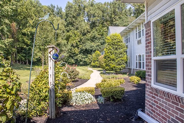 A landscaped outdoor pathway beside a building with white siding and brick accents, surrounded by green bushes, trees, and garden decorations under a clear sky.