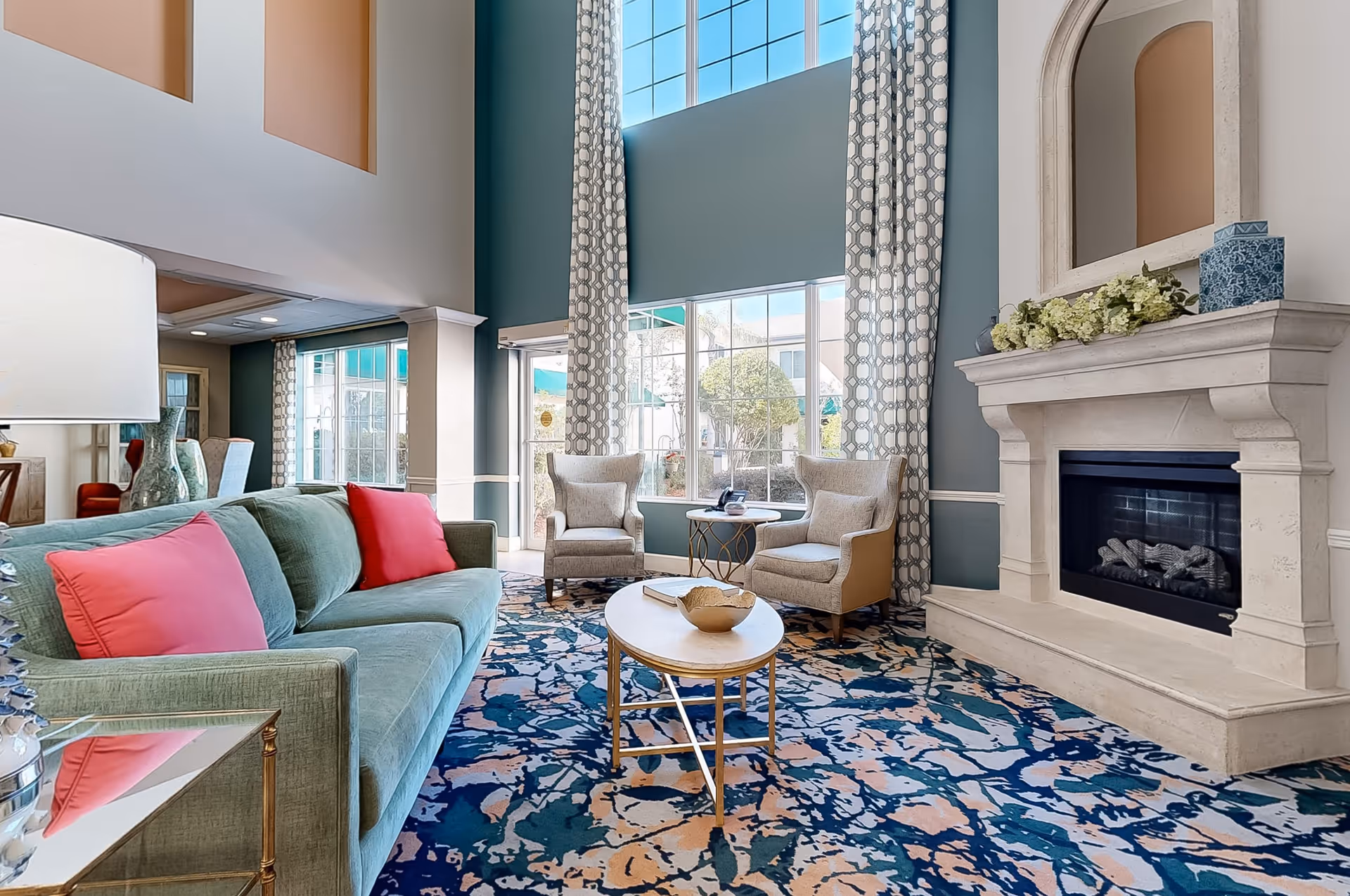 Bright and spacious living room with a high ceiling, large windows with patterned curtains, a green sofa with pink cushions, two beige armchairs, a decorative fireplace with a mantel adorned with flowers and a blue vase, and a colorful patterned carpet.