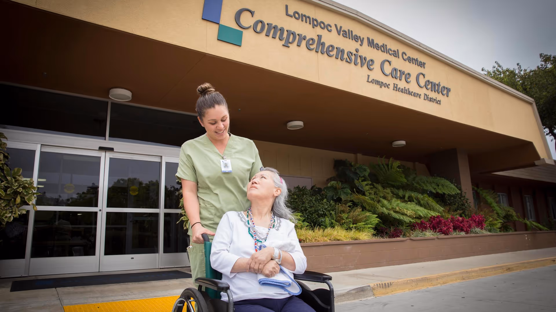 A healthcare worker in green scrubs is pushing an elderly woman in a wheelchair outside the entrance of Lompoc Valley Medical Center Comprehensive Care Center. The elderly woman is looking up at the healthcare worker, and there are plants and shrubs along the building exterior.