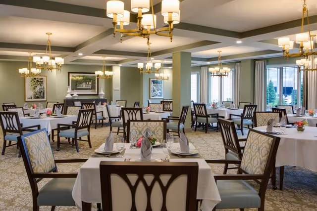 A spacious and elegant dining room with multiple tables covered in white tablecloths, each set with plates, glasses, and folded napkins. The room features patterned carpet, large windows with curtains, framed artwork on the walls, and several gold chandeliers providing warm lighting.