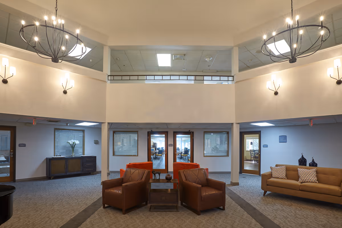 A spacious senior living facility common area with two brown leather armchairs and a small wooden table between them in the center. To the right is a brown sofa with two patterned pillows and decorative vases on a side table. The room features beige carpet, white walls, and multiple light fixtures including wall sconces and two large circular chandeliers hanging from the ceiling. There are windows and doors leading to other rooms in the background.