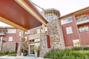 Exterior view of a multi-story senior living facility with red siding and stone accents, featuring a covered entrance and balconies on the upper floors, surrounded by greenery.