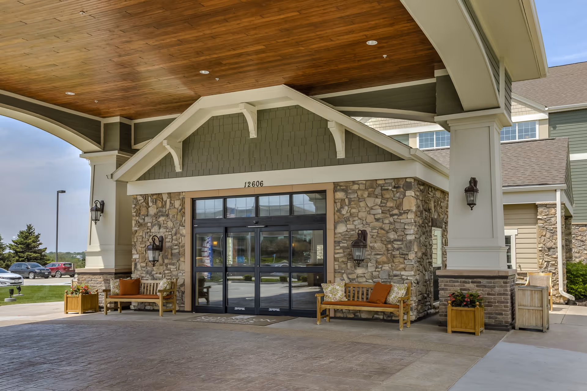 Entrance of a retirement community building with stone and siding exterior, automatic glass doors, wooden benches with cushions on either side, potted plants, and a covered driveway with a wooden ceiling.
