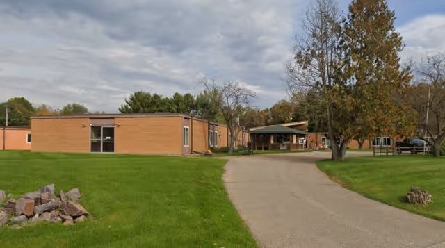 Exterior view of a single-story brick building with a paved driveway leading to a covered entrance. The building is surrounded by green grass, trees with autumn foliage, and a partly cloudy sky.