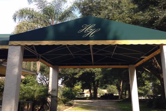 Green entrance canopy with gold trim and a scripted "HH" monogram over a driveway supported by four columns and surrounded by trees.