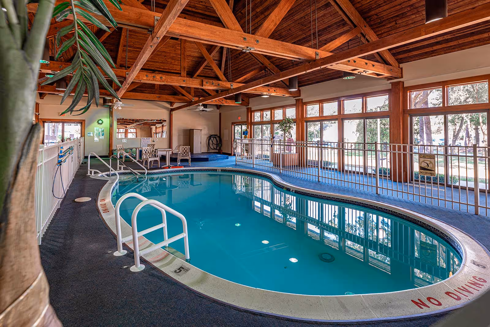 Indoor swimming pool in a wood‑beamed community room with large windows, railings, and seating.