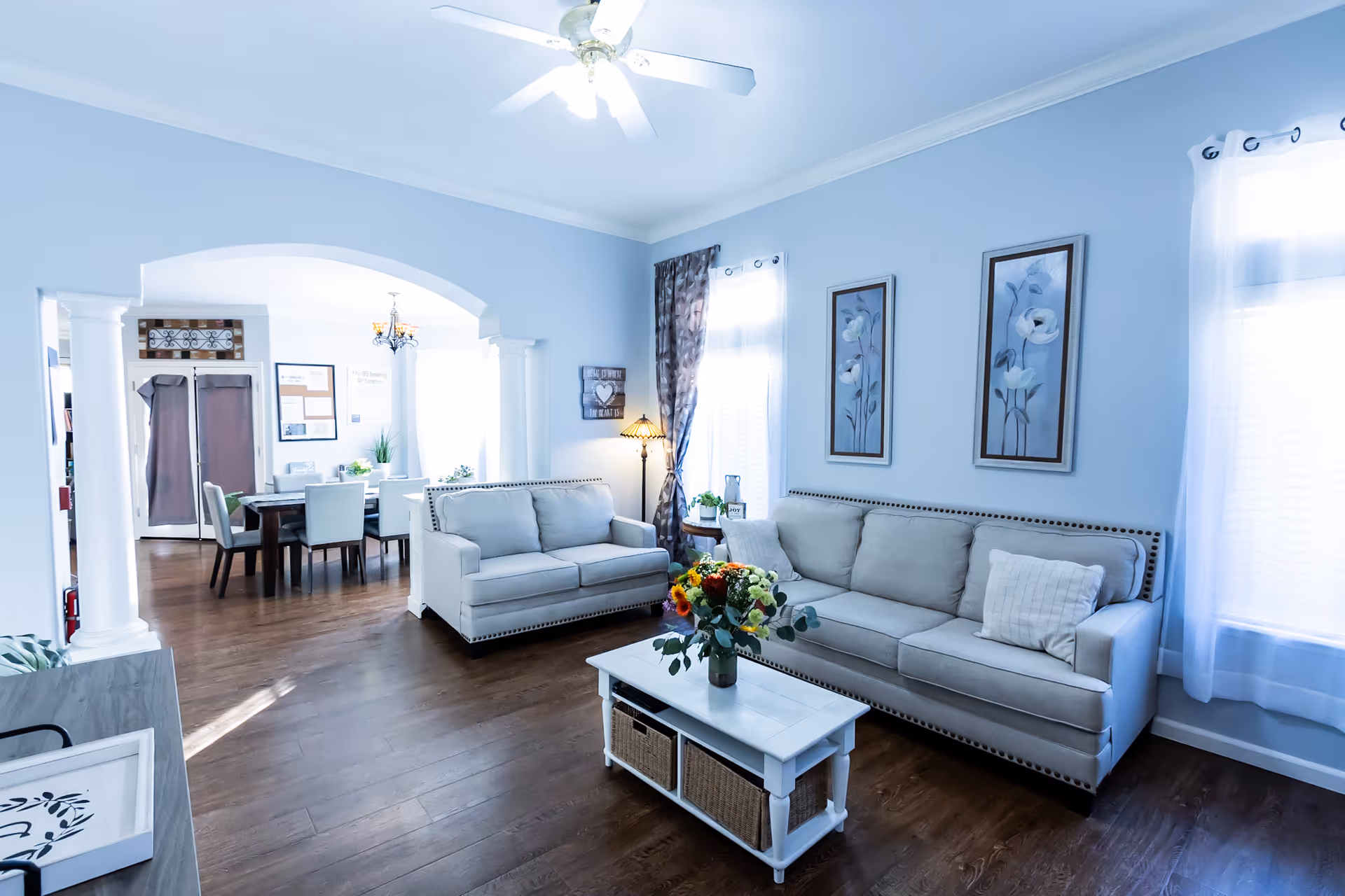A bright and airy living room with light blue walls, two beige sofas, a white coffee table with a flower vase, and two framed floral paintings on the wall. The room has large windows with sheer white curtains and a ceiling fan. In the background, a dining area with a table and chairs is visible through an arched opening supported by white columns.
