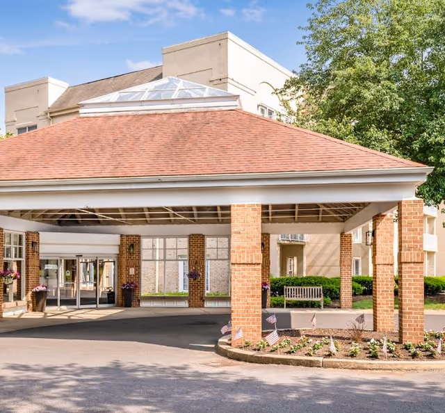 Entrance area of a senior living facility with a covered drop-off zone supported by brick pillars, a flower bed with small American flags, and a bench near the building. The building has large windows and a beige exterior with a red roof over the entrance.