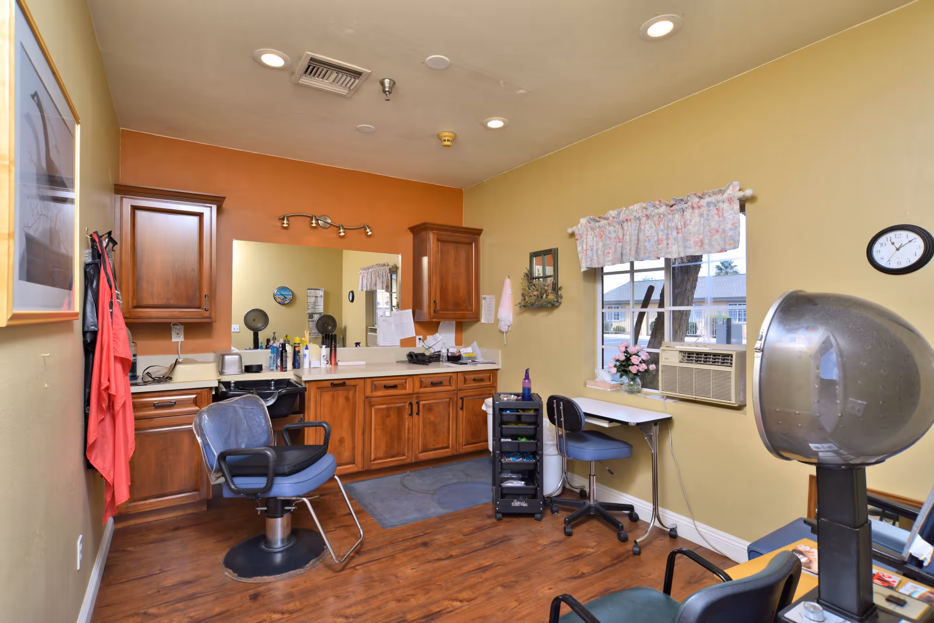 Interior view of a senior living facility's hair salon area with wooden cabinets, a large mirror, salon chairs, a hair dryer, and a small desk with a chair near a window with floral curtains. The room has yellow and orange walls and wood flooring.