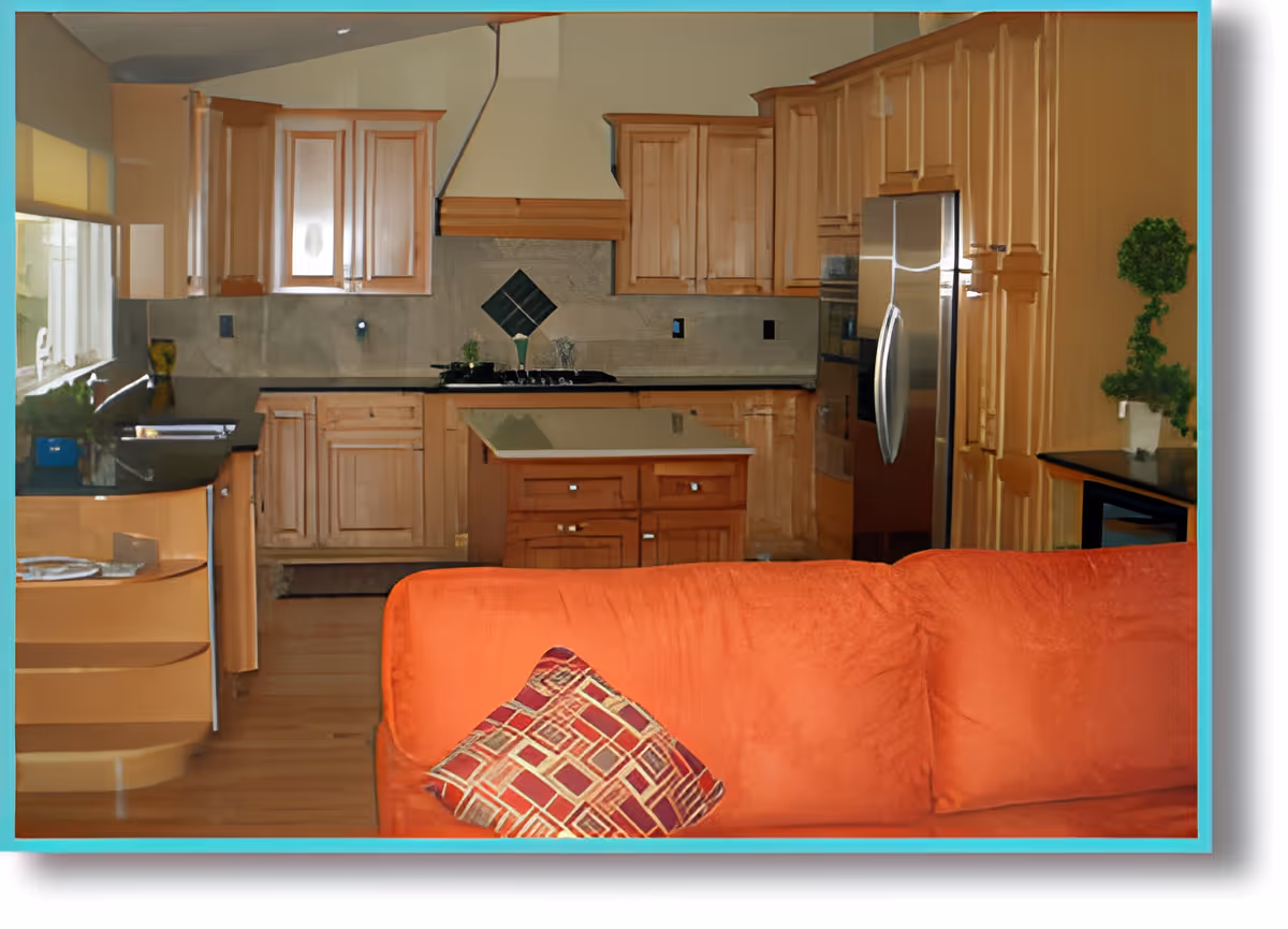 Interior view of a kitchen with wooden cabinets, a central island, stainless steel refrigerator, and a black countertop. In the foreground, there is an orange couch with a patterned cushion. A small potted plant is on the right countertop.