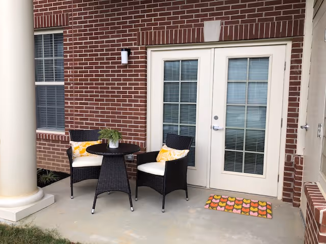 A small covered brick patio with two wicker chairs, a round table, and French doors.