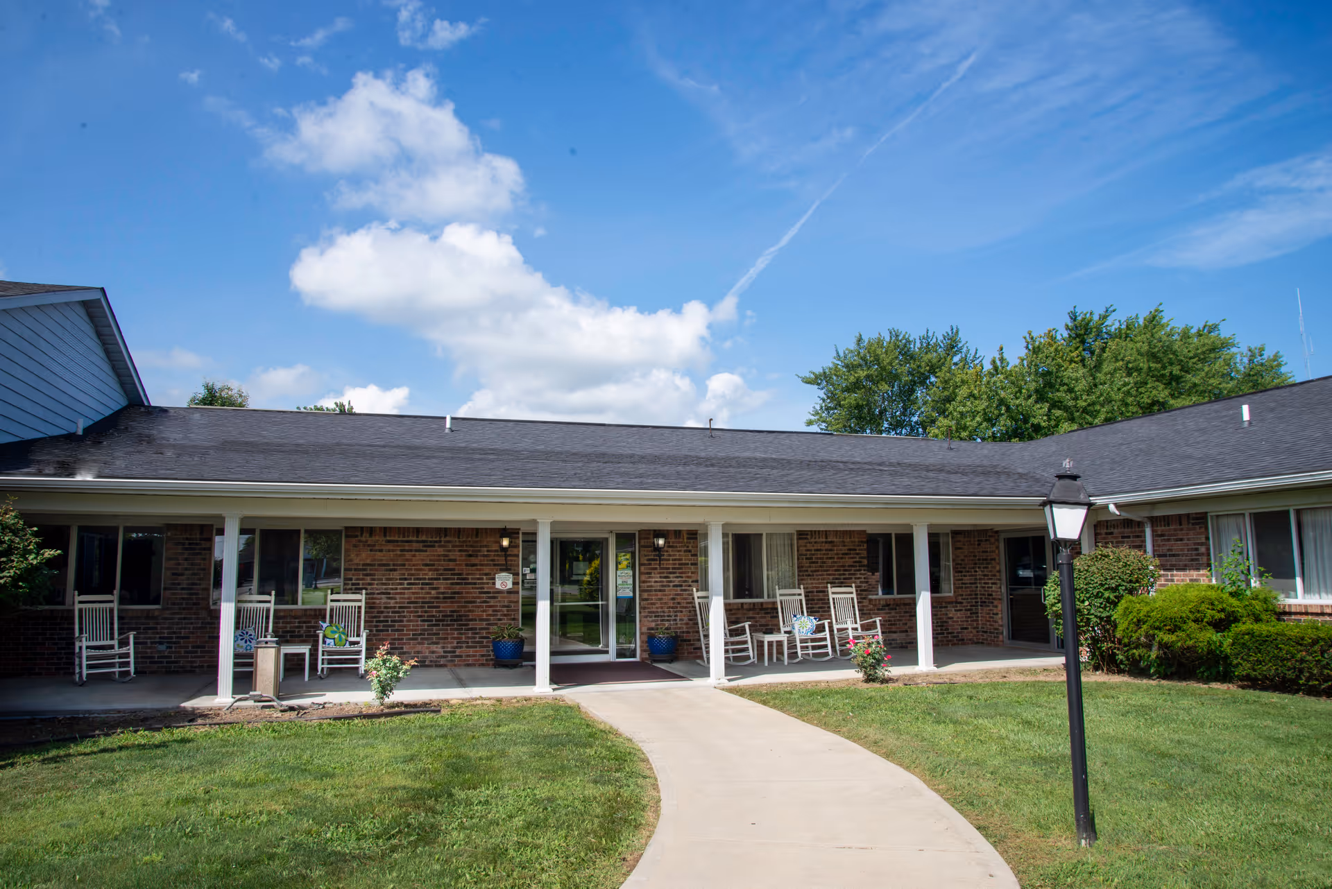 Front exterior view of Manderley Health Care Center showing a single-story brick building with a covered porch, several white rocking chairs, potted plants, a lamp post, and a curved concrete walkway leading to the entrance under a blue sky with some clouds.