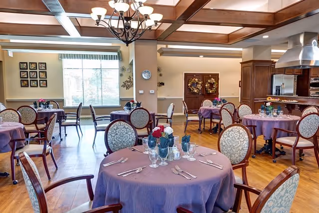 A dining room with round tables covered in purple tablecloths, each set with glassware, silverware, and floral centerpieces. The room features wooden floors, upholstered chairs with patterned backs, a chandelier hanging from a coffered ceiling, and a kitchen area with stainless steel appliances in the background.