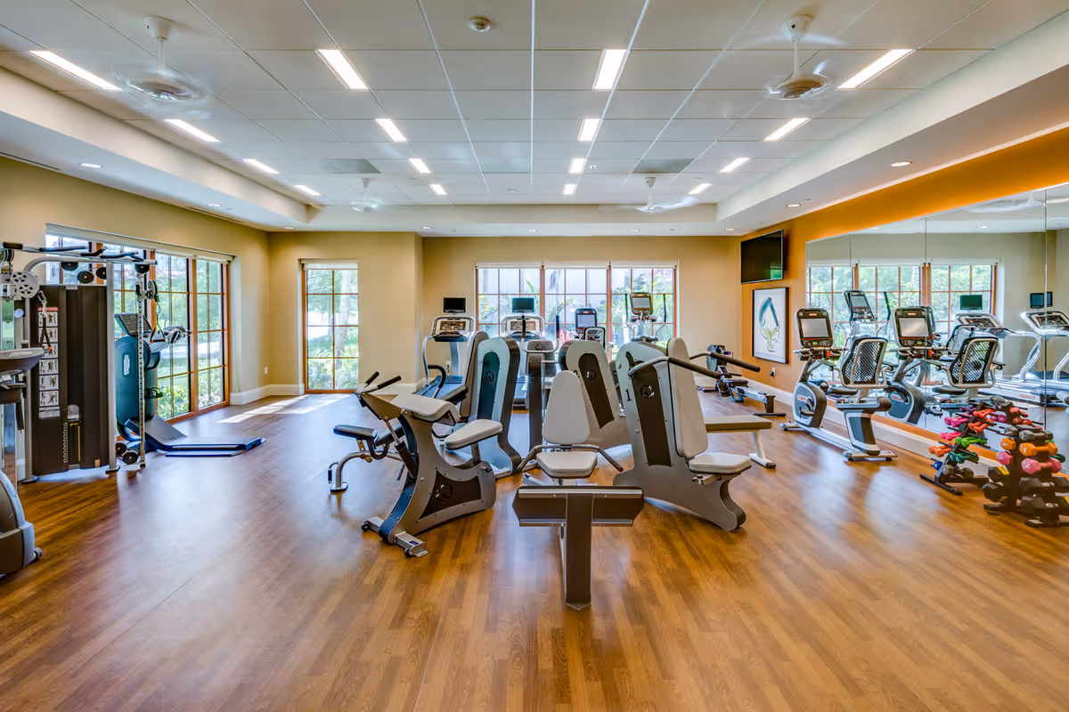A bright and spacious fitness room with various exercise machines including weight machines, stationary bikes, and treadmills. The room has wooden flooring, large windows letting in natural light, ceiling fans, and a wall covered with mirrors. There is also a rack of colorful dumbbells on the right side.