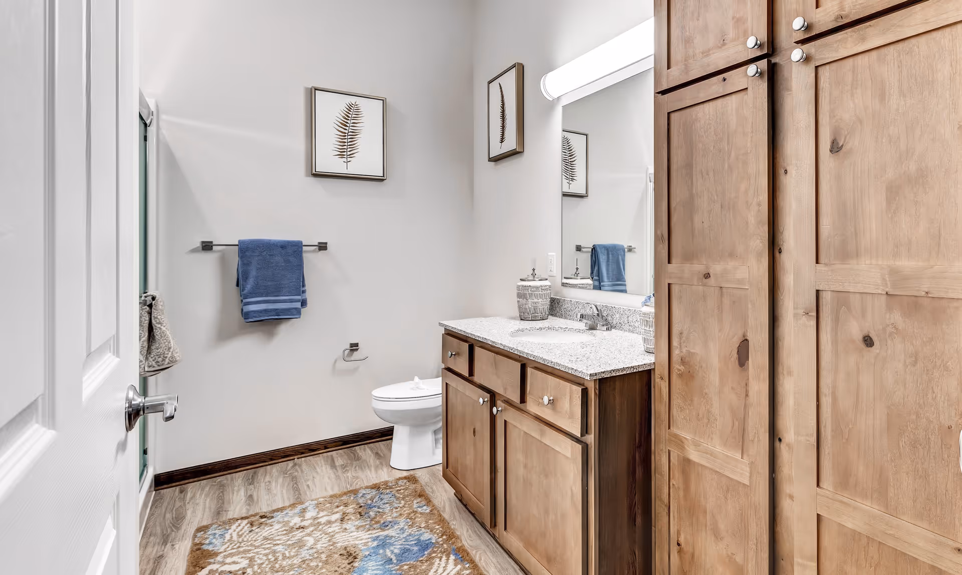A bathroom with wooden cabinets, a granite countertop with a sink, a large mirror with a light above it, a toilet, a towel rack with a blue towel, a framed leaf artwork on the wall, and a patterned rug on the wooden floor.
