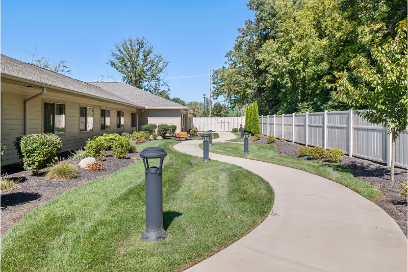 Curved concrete pathway lined with black lamp posts and green grass, leading past a beige building with windows on the left and a white fence on the right, surrounded by trees and shrubs under a clear blue sky.