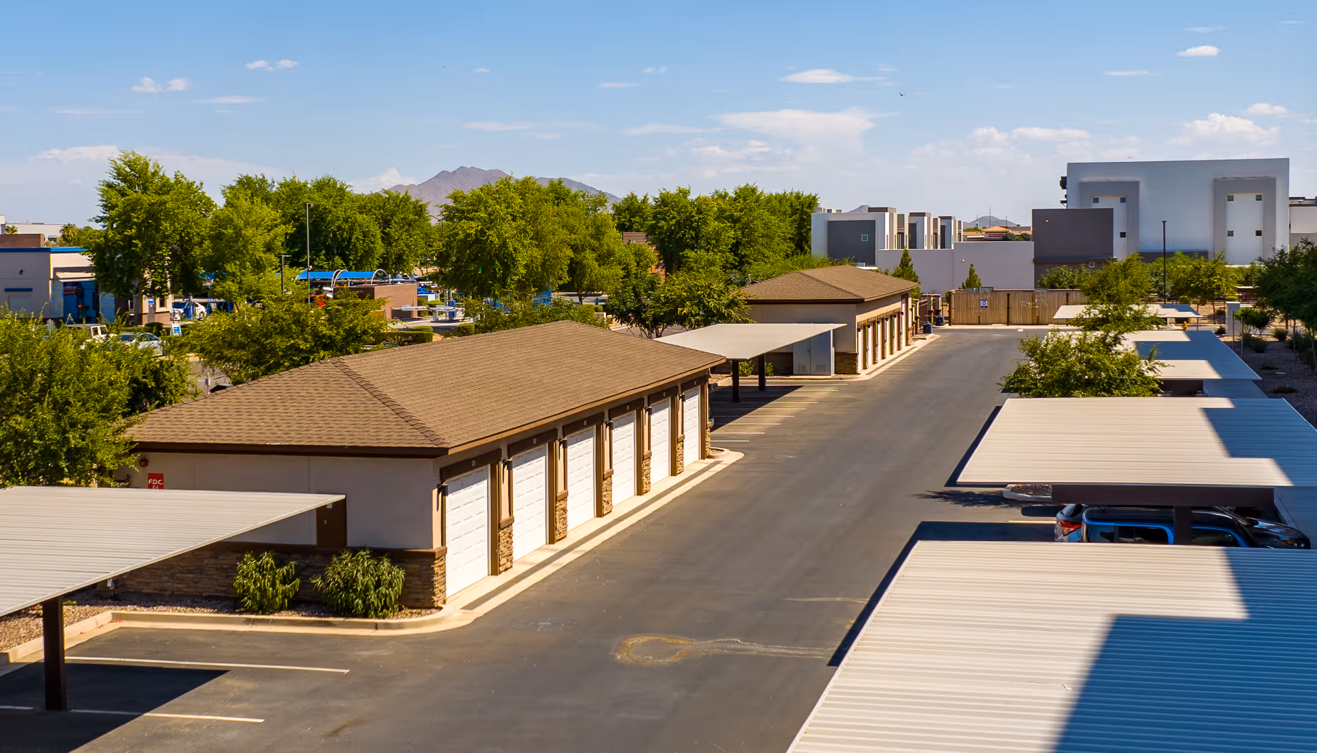 View of a parking area with covered parking spaces and two long buildings with multiple garage doors, surrounded by trees and other buildings in the background under a clear blue sky.
