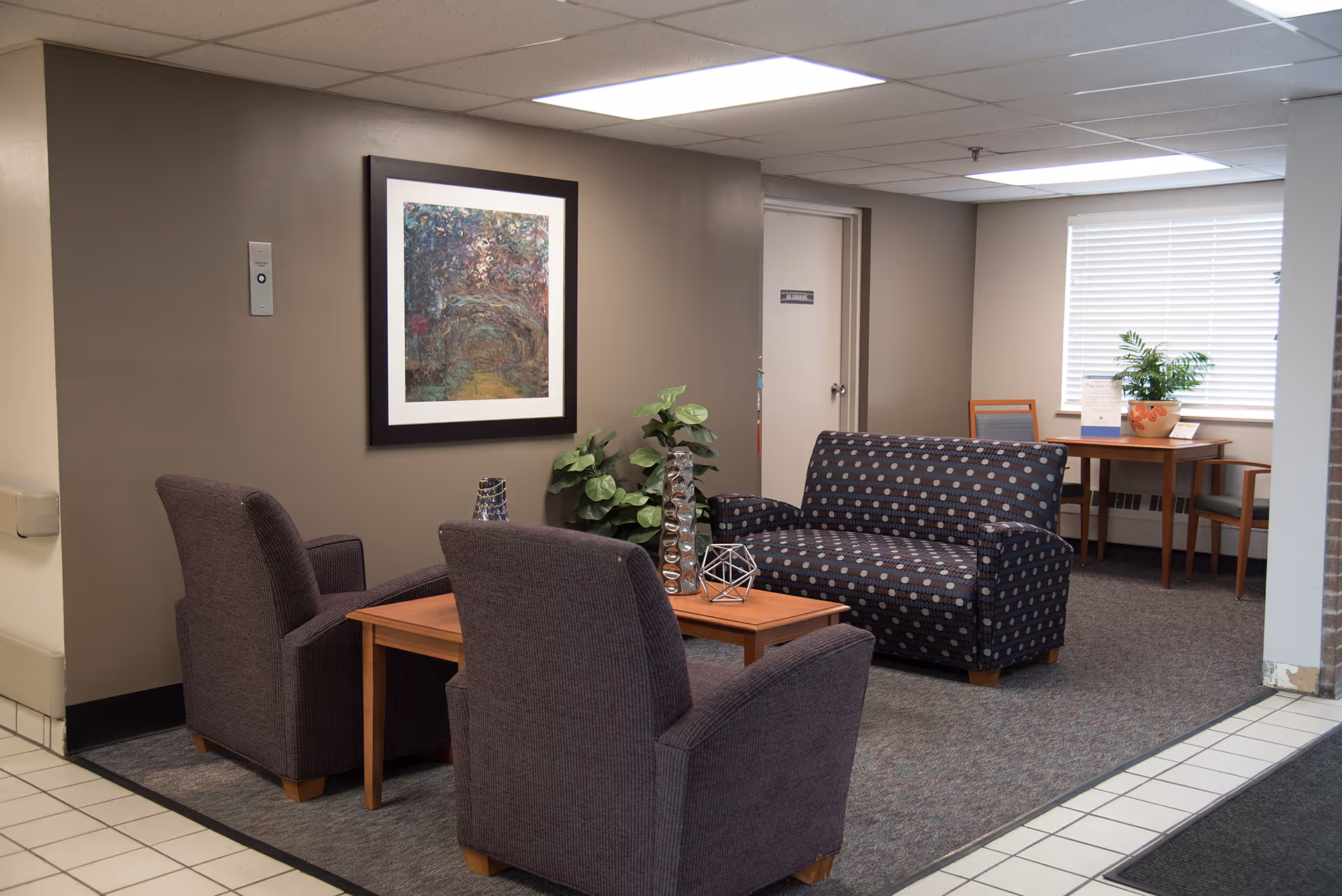A cozy seating area in a senior living facility with two upholstered armchairs and a patterned loveseat arranged around a wooden coffee table. Decorative items and a potted plant are on the table. A framed abstract painting hangs on the wall, and a window with blinds lets in natural light. In the background, there is a small table with chairs and another potted plant.