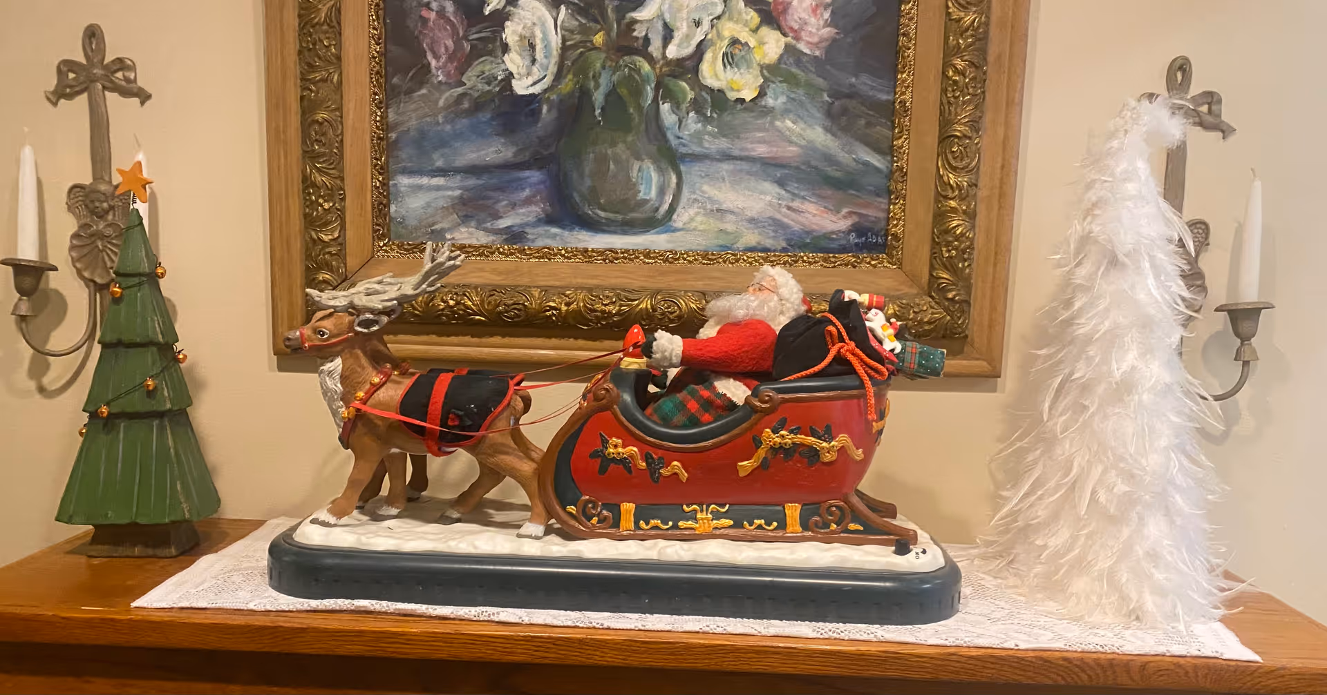 A decorative Christmas display on a wooden surface featuring a figurine of Santa Claus in a red sleigh pulled by a reindeer, flanked by a green wooden Christmas tree with a star on top on the left and a white feathered Christmas tree on the right. Behind the display is a framed painting of flowers in a vase and two wall-mounted candle holders with white candles.