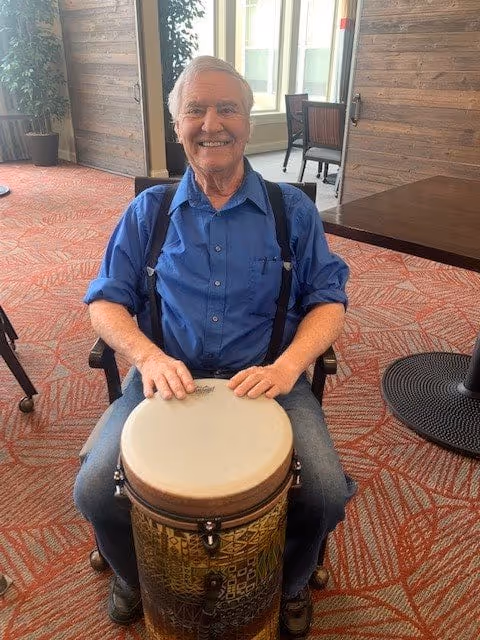 An elderly man wearing a blue shirt and suspenders is sitting on a chair indoors, smiling and playing a drum. The room has a patterned carpet, wooden walls, and large windows letting in natural light.