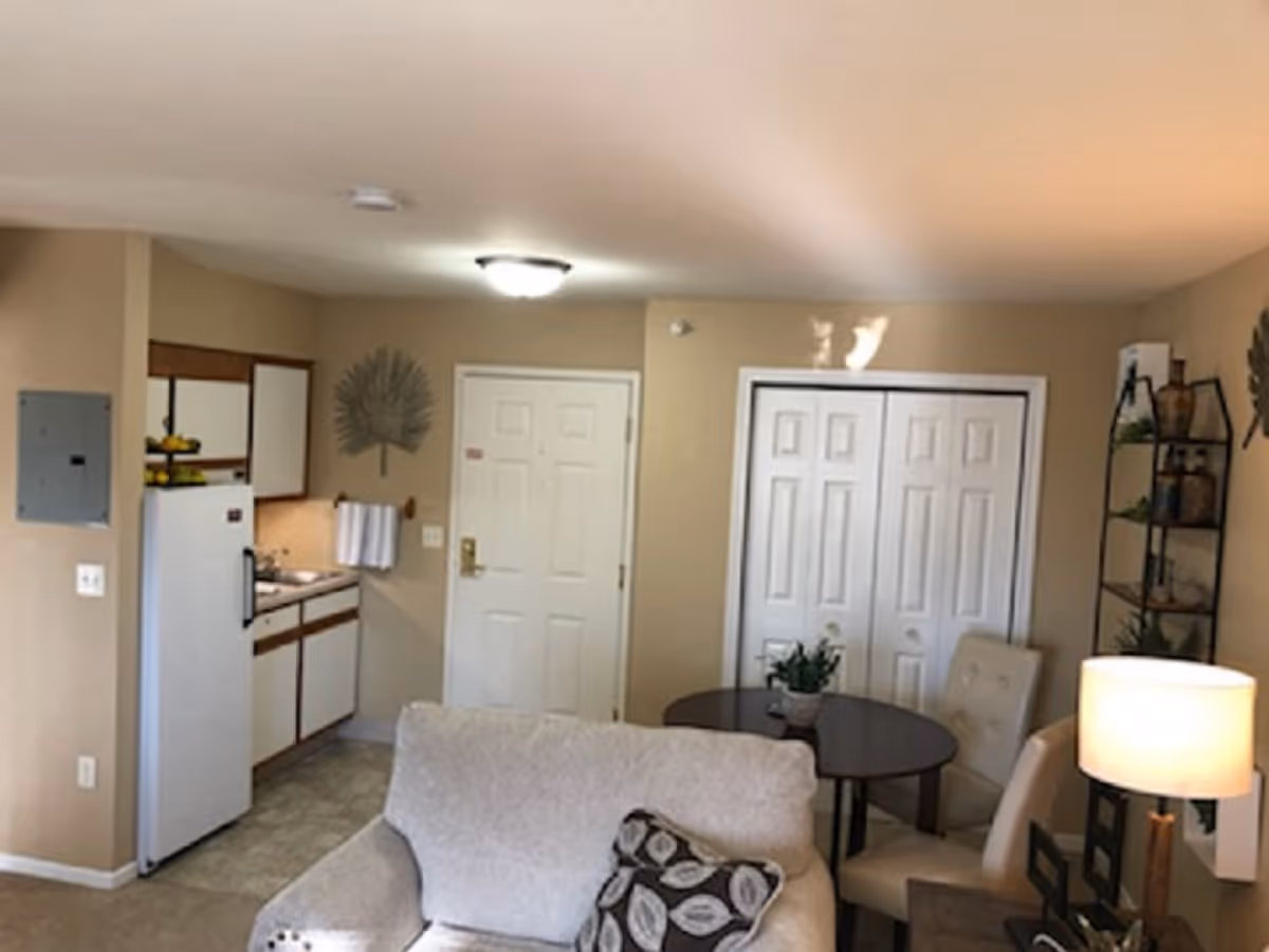 Interior view of a senior living facility apartment showing a small kitchen area with white cabinets and a refrigerator on the left, a beige armchair with a patterned pillow in the foreground, a round dining table with two chairs near a set of white double doors, and a floor lamp on the right side. The walls are painted beige and decorated with a metal wall art piece and a shelving unit with decorative items.