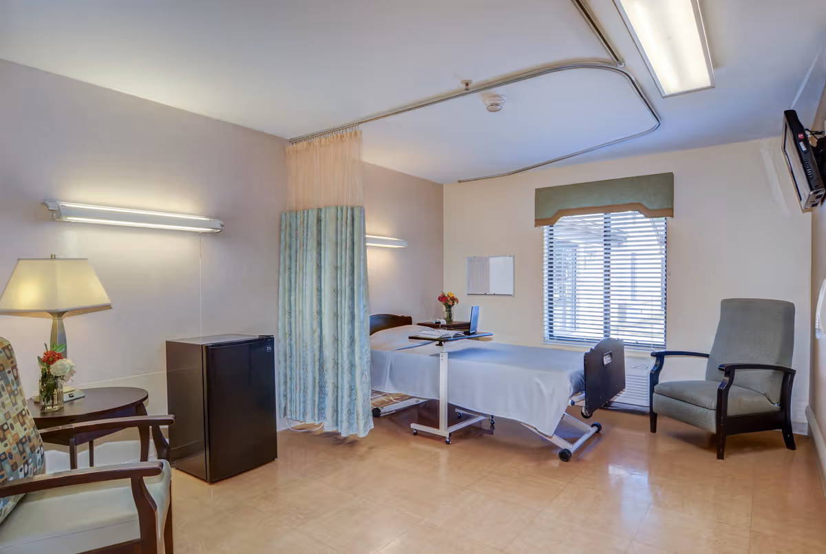 A senior care patient room with a hospital bed, privacy curtain, chairs, a small refrigerator, and a window with blinds.