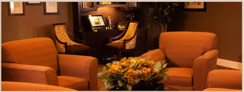 A cozy living room area with three orange upholstered armchairs arranged around a table with a floral centerpiece. In the background, there is a dark wooden desk with a computer and two patterned chairs. The room has warm lighting and brown walls.