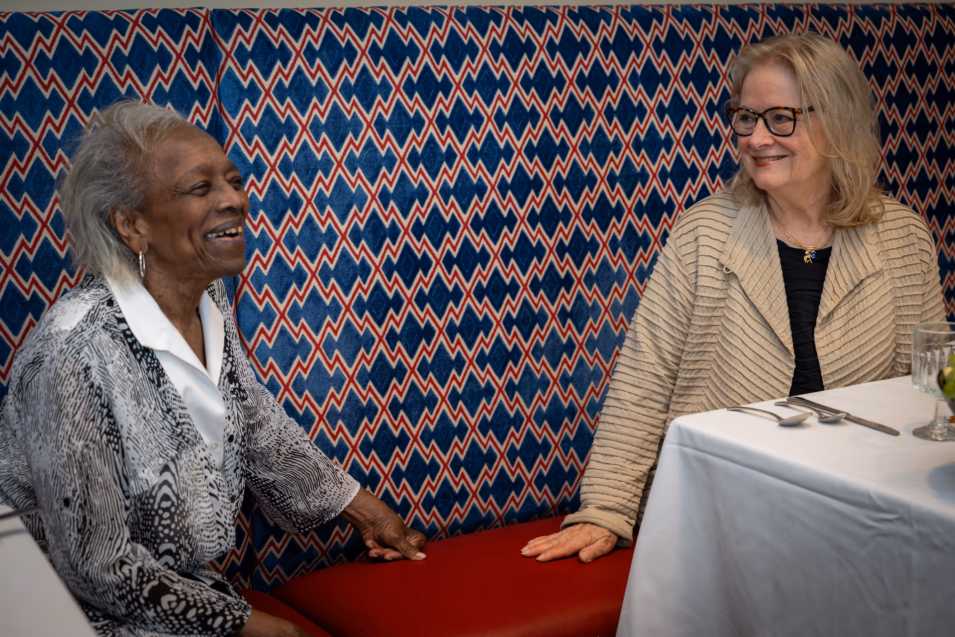 Two elderly women smiling and talking while seated in a patterned booth at a dining table set with utensils and a glass.