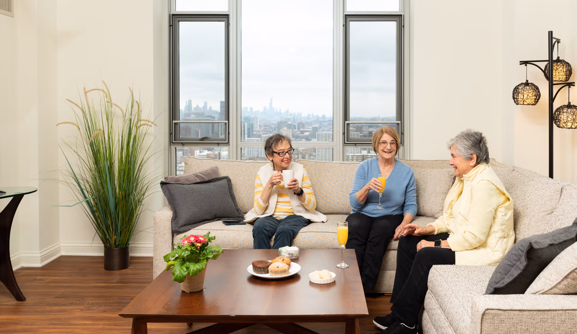 Three elderly women sitting on a beige sectional sofa in a bright living room with large windows showing a cityscape view. They are smiling and holding drinks, with a wooden coffee table in front of them holding a potted plant, muffins, and other small dishes. The room has wooden flooring, a tall green plant in a pot, and a floor lamp with three decorative lights.