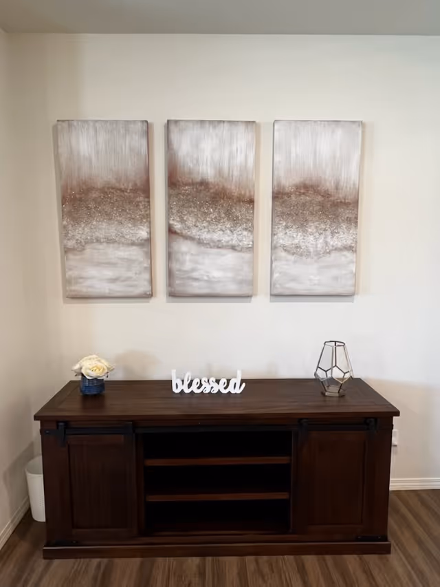 A dark wooden cabinet with sliding doors and open shelves in the center, placed against a white wall. On top of the cabinet, there is a small blue vase with white flowers on the left, a decorative word sign that says 'blessed' in the middle, and a geometric candle holder on the right. Above the cabinet, three abstract paintings with shades of brown, beige, and white are hung side by side.