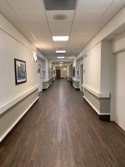A long, clean hallway in a senior living facility with wood-look flooring, white walls, handrails on both sides, framed artwork, and ceiling lights evenly spaced along the corridor.