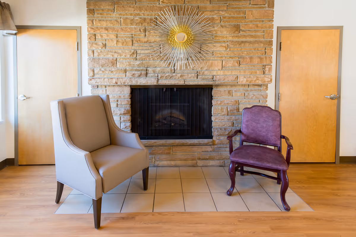 Two chairs placed in front of a stone fireplace with a sunburst mirror and two wooden doors on either side.