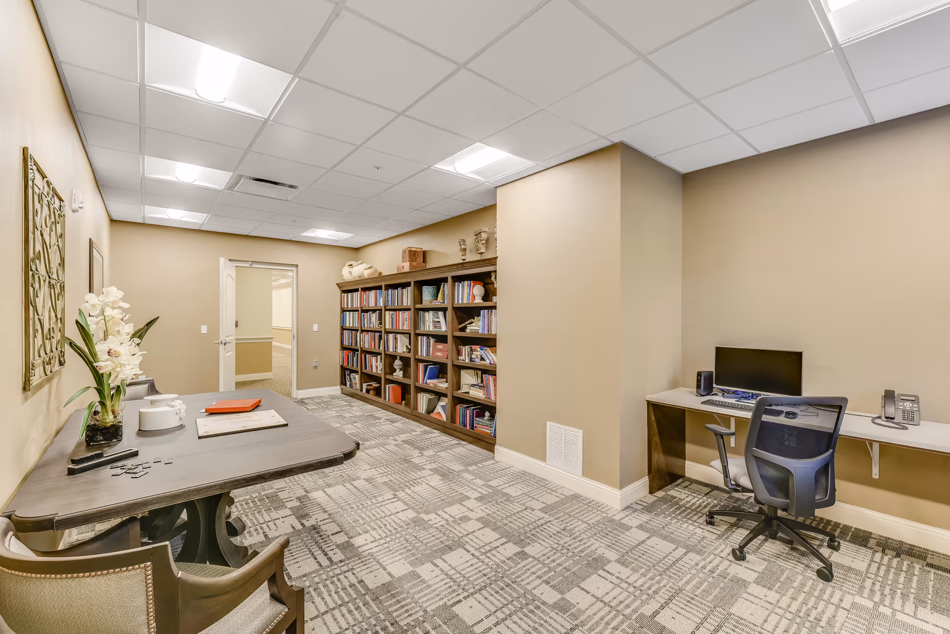 Interior view of a senior living facility room with beige walls and patterned carpet. The room features a long wooden table with chairs on the left side, decorated with a plant and some papers. On the right side, there is a built-in desk with a computer, office chair, and telephone. At the back, there is a large bookshelf filled with books and decorative items. The ceiling has recessed lighting panels.