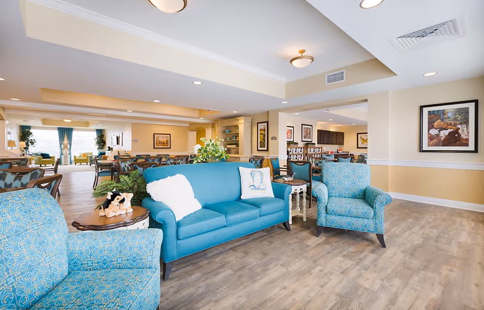 A bright and spacious common area in a senior living facility featuring a teal sofa with white and patterned pillows, two matching patterned armchairs, wooden tables, and a dining area with multiple tables and chairs in the background. The room has light-colored walls, wood flooring, framed artwork on the walls, and large windows with blue curtains allowing natural light to fill the space.