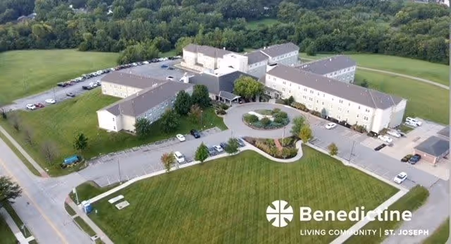 Aerial view of Benedictine Living Community-St. Joseph, showing a large multi-wing building surrounded by green lawns, trees, parking lots with several cars, and nearby roads.