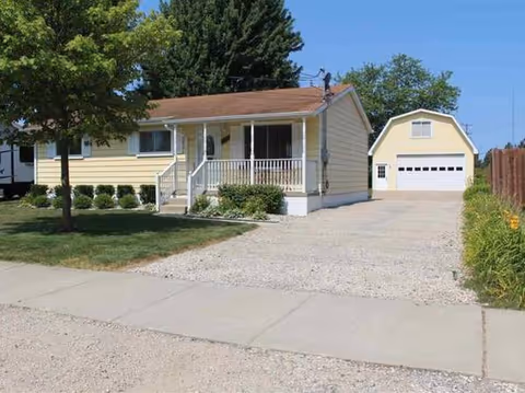 A small beige single-story house with a front porch, gravel driveway, and detached garage.