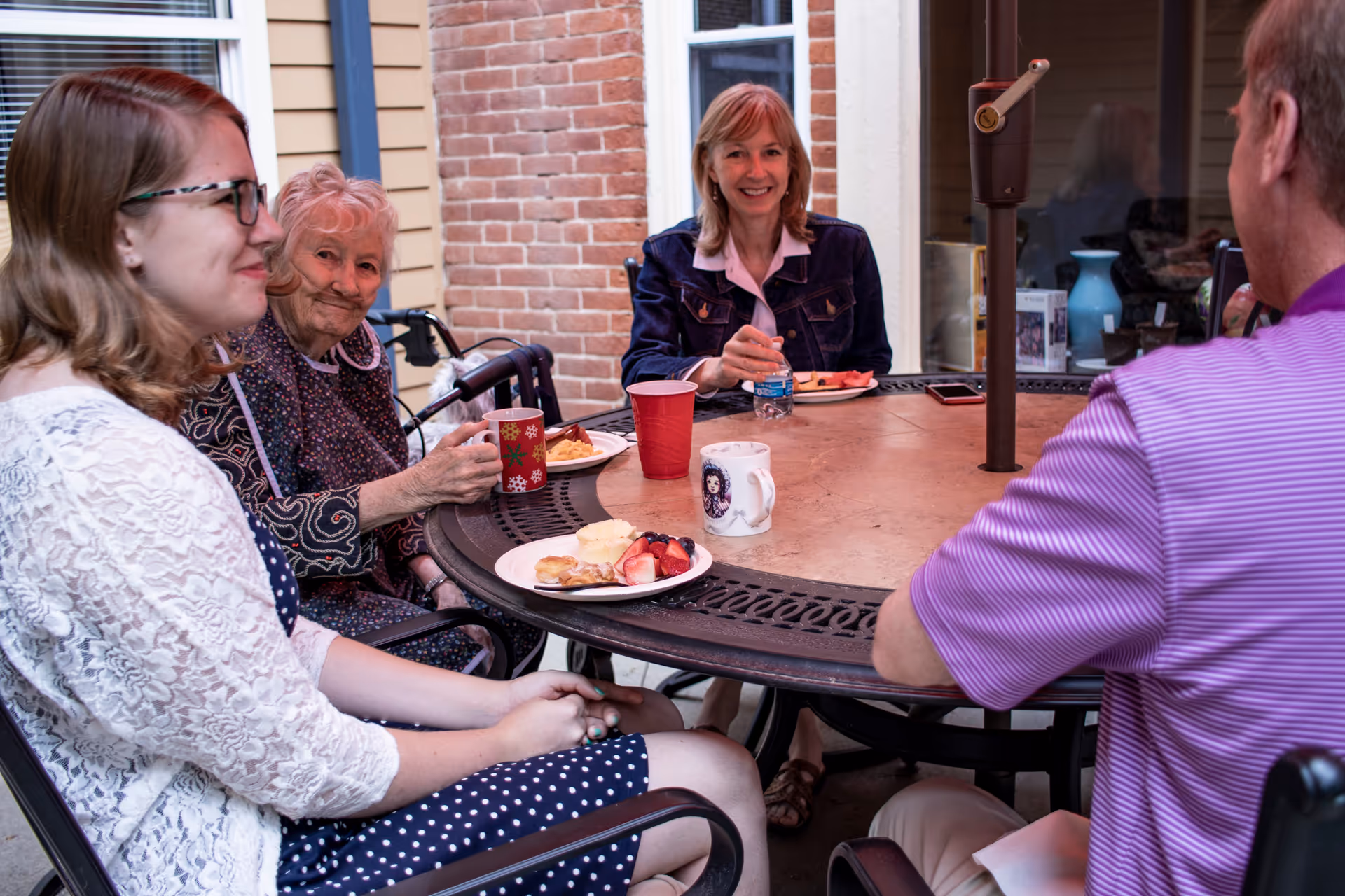 Four people sitting around an outdoor round table enjoying a meal together. The group includes an elderly woman with a nasal cannula, a younger woman in glasses and a white lace cardigan, a middle-aged woman in a denim jacket, and a man in a purple striped shirt. Plates with food and cups are on the table, and the setting appears to be a patio area with brick and siding walls in the background.