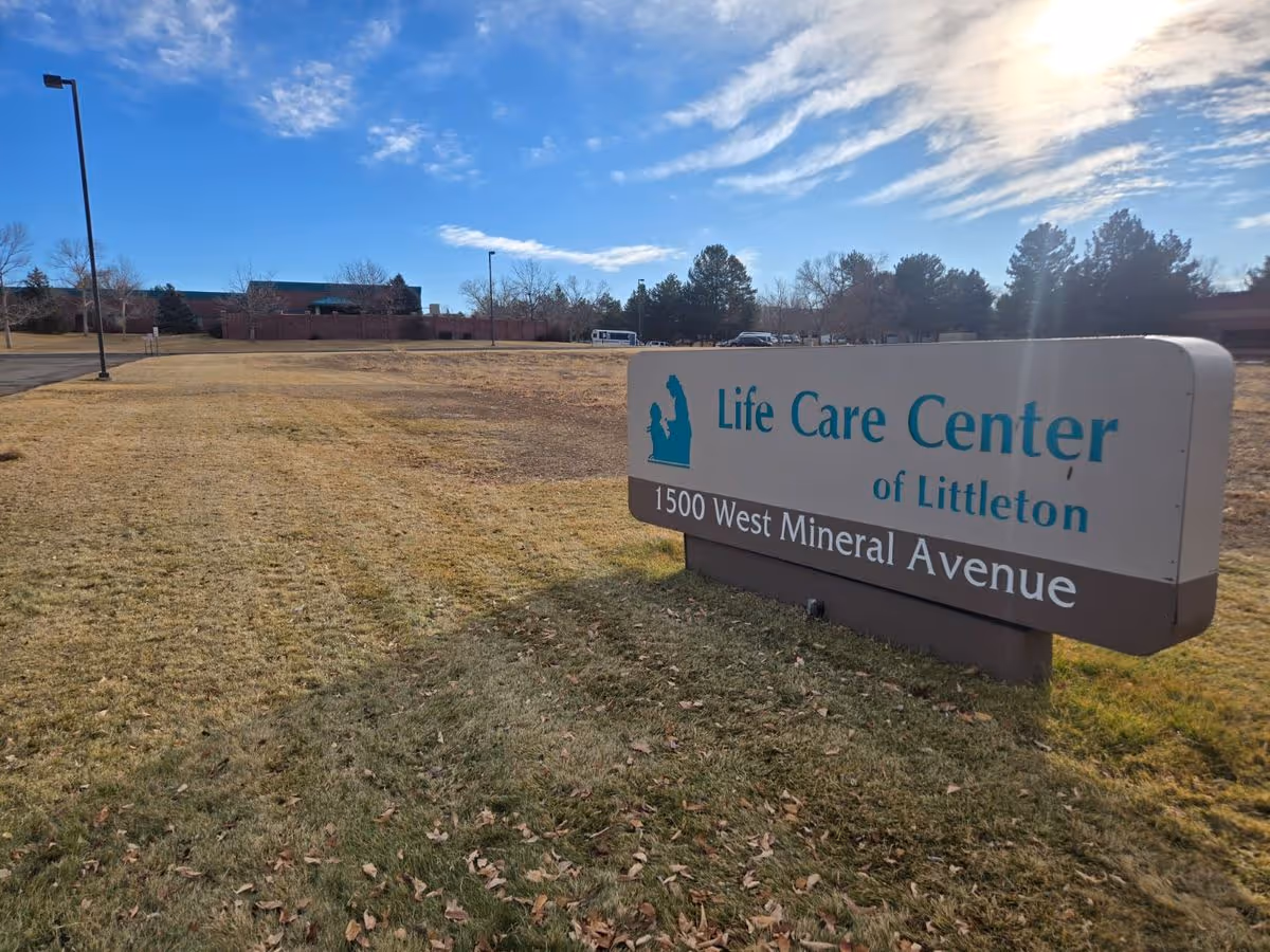 Outdoor sign for Life Care Center of Littleton on a grassy lawn with the facility buildings and a blue sky in the background.
