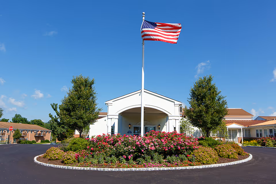 Front exterior view of Fort Washington Estates building with a circular driveway, landscaped flower beds, two trees, and an American flag on a flagpole in the center under a clear blue sky.