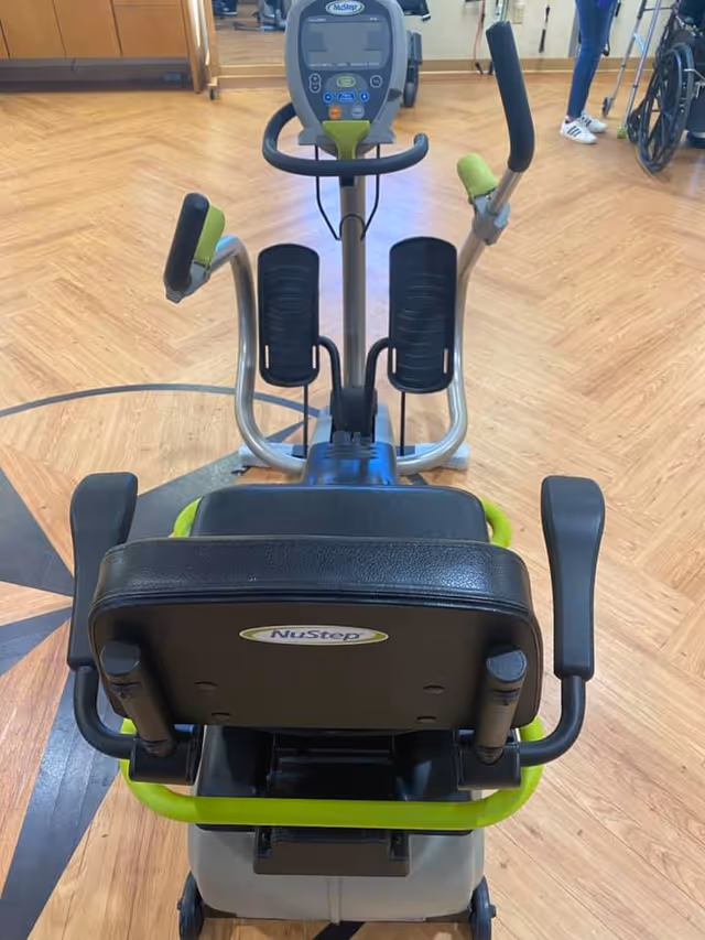 A NuStep exercise machine positioned on a wooden floor in a room with mirrored walls. Part of a wheelchair and a person wearing blue pants and white sneakers are visible in the background.