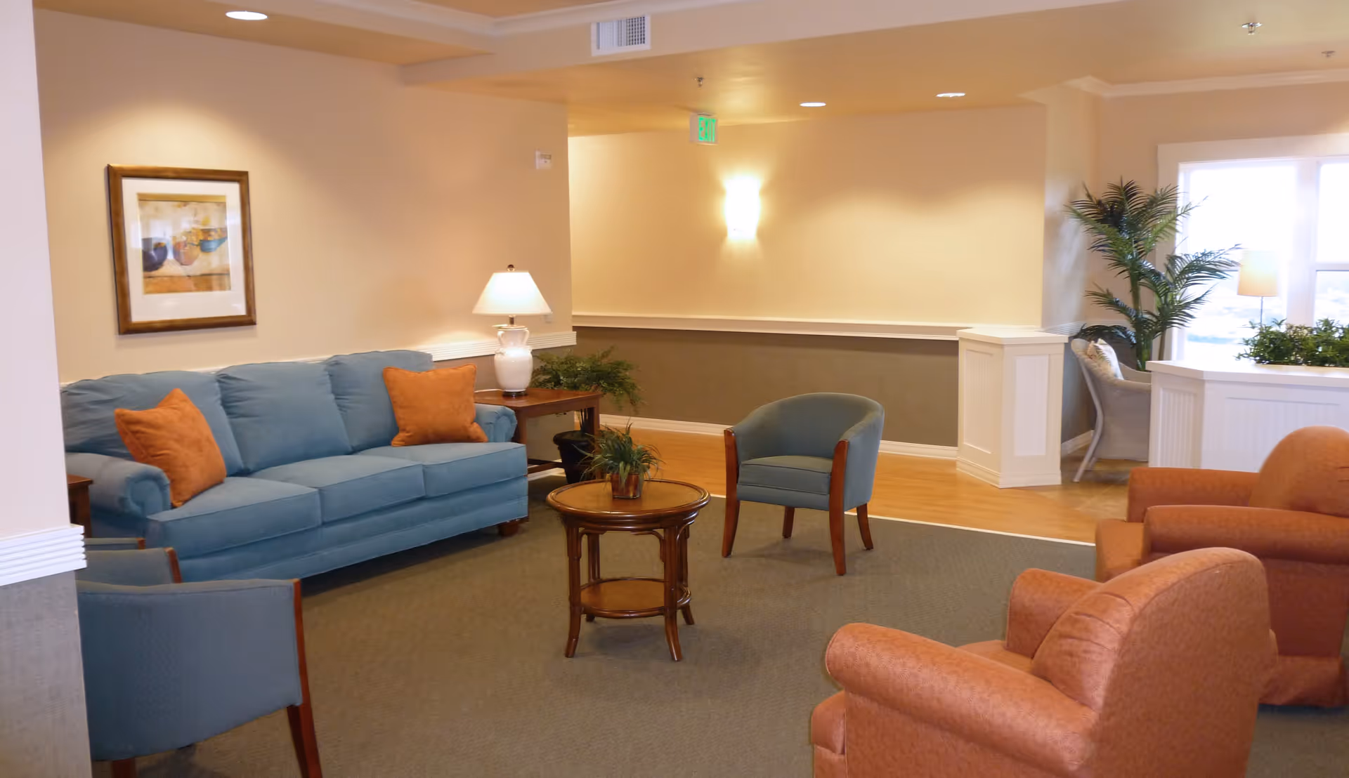 A cozy living room area with a blue sofa adorned with orange pillows, two blue armchairs, and two orange armchairs arranged around a small round wooden coffee table with a plant on it. There is a side table with a white lamp and a framed picture on the wall. The room has warm lighting, beige walls, and a large window letting in natural light with plants nearby.