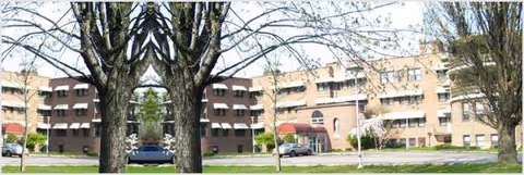 A brick senior living building with arches, trees, and parked cars in the foreground.