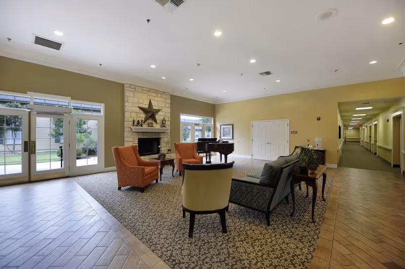 Spacious senior living lounge with chairs, a sofa, a fireplace, and a piano near large windows and doors.